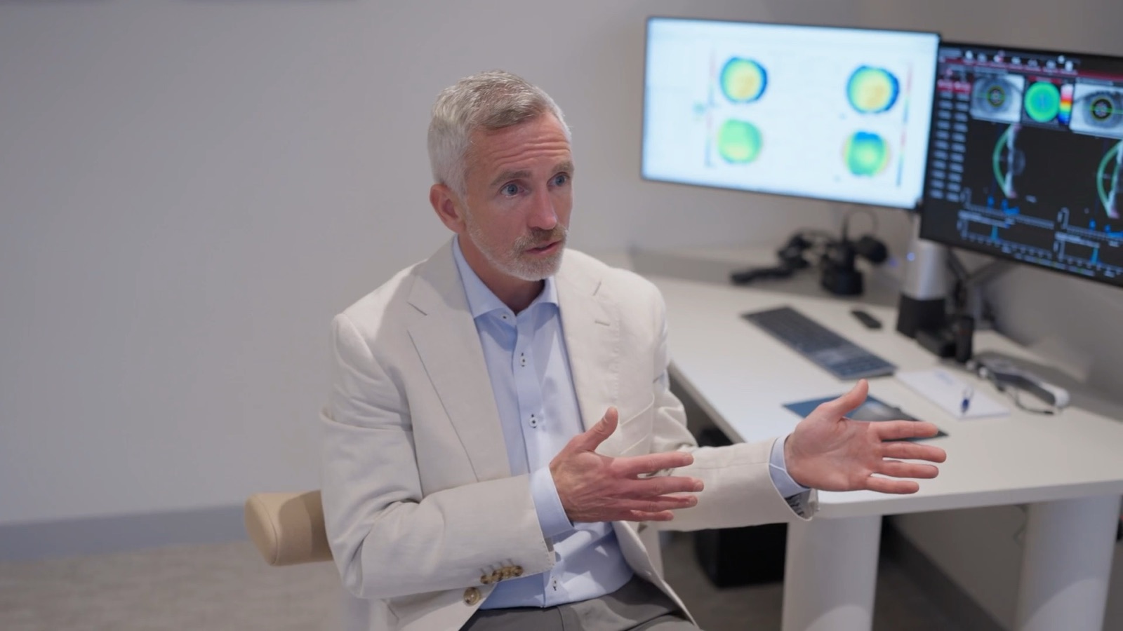 Dr Richard Barry in a consulting room with corneal mapping and eye scans on screen